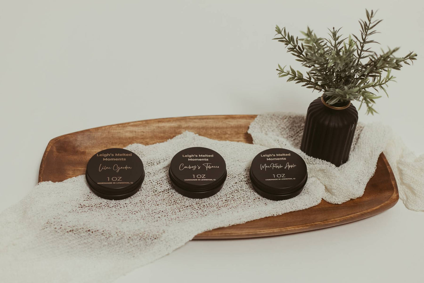 Three 1oz Leigh's Melted Moments wax melt containers on a wooden tray with a decorative plant.