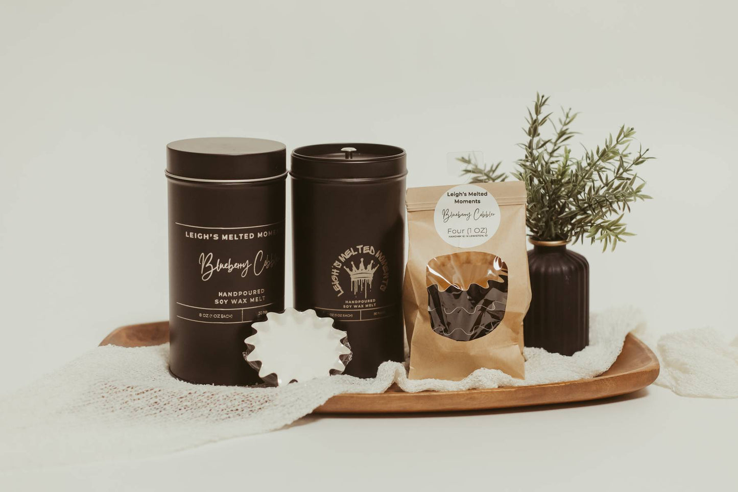 Leigh's Melted Moments soy wax melts in black tins and kraft packaging on a wooden tray with a decorative plant.