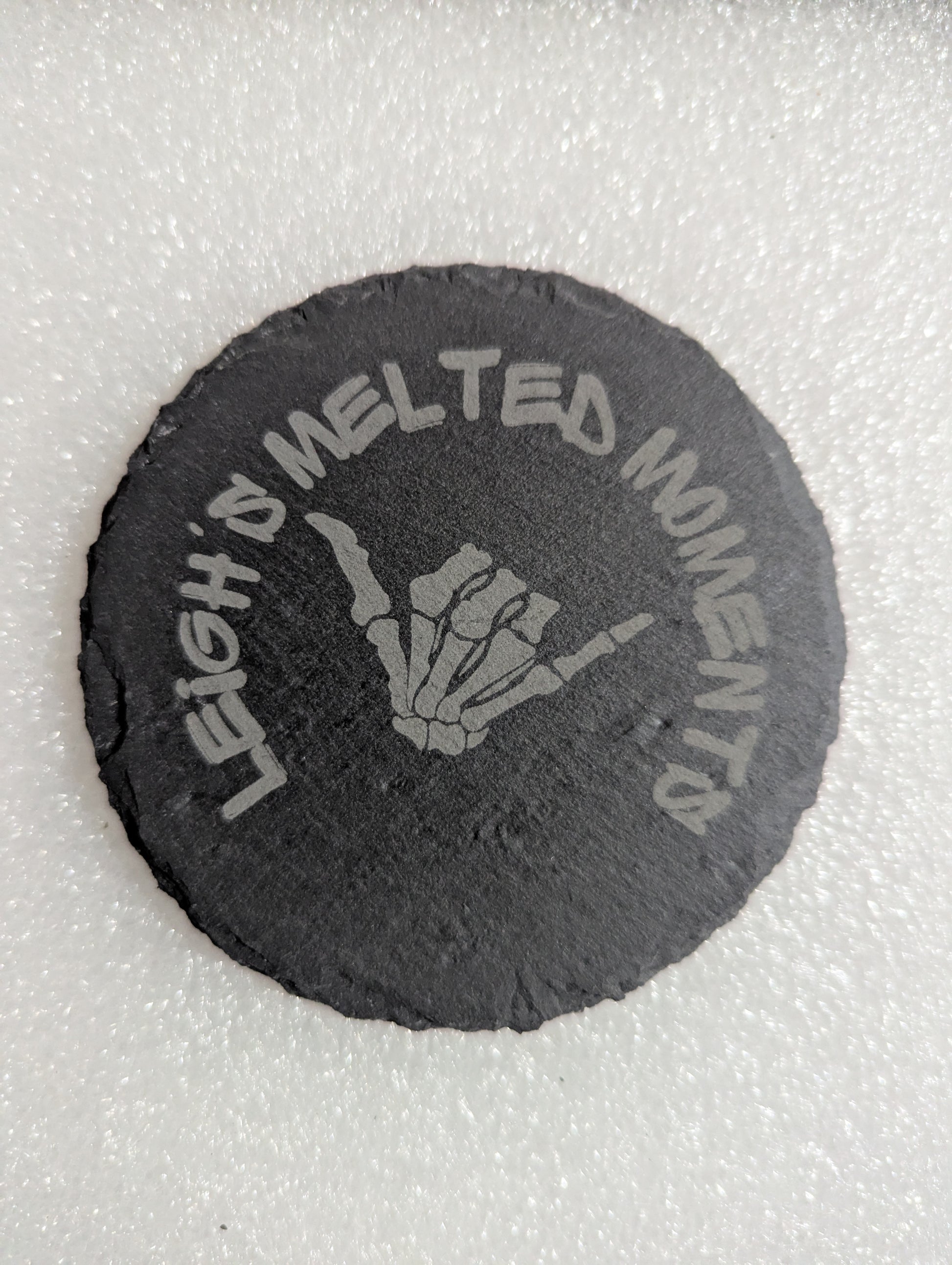 Round slate coaster with an engraved skeletal hand making a gesture, accompanied by 'Leigh's Melted Moments' branding.